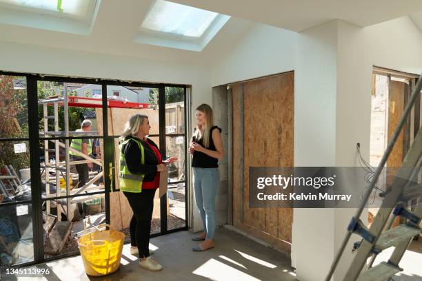 a female  builder stands in a building site amongst a half finished renovation with bare wooden cupboards half finished. she talks through the project with her client - home inspection photos et images de collection