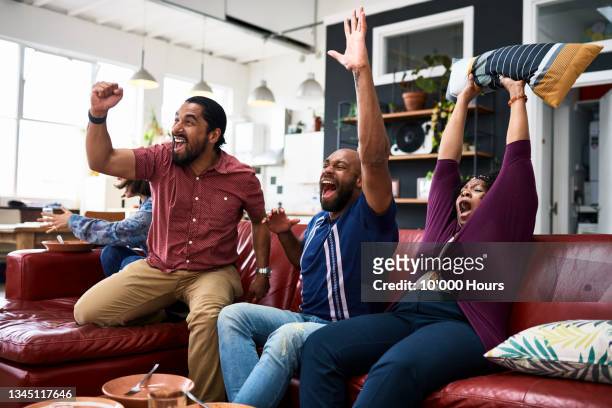 friends watching tv at home and cheering with arms raised - friendly match photos et images de collection