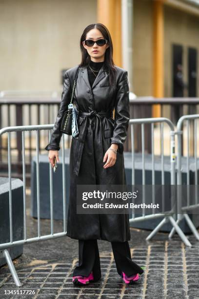 Model wears black sunglasses from Chanel, gold earrings, a black wool turtleneck pullover, gold chain necklaces, a black shiny leather belted trench...