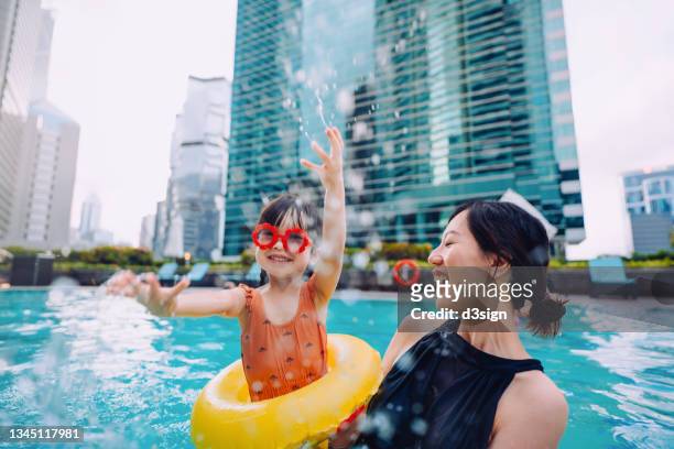 Happy little Asian girl with sunglasses smiling joyfully, splashing and playing with water with mother, enjoying family bonding time in the swimming pool on Summer vacation