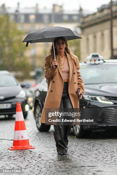 Guest wears a black umbrella, gold chain pendant necklaces from Chanel, beige ribbed cropped t-shirt, a beige long oversized coat, a gold Juste Un...