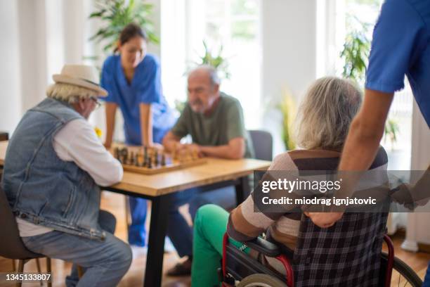 senior man in wheelchair watching his friends playing chess at nursing home - nursing assistant stock pictures, royalty-free photos & images