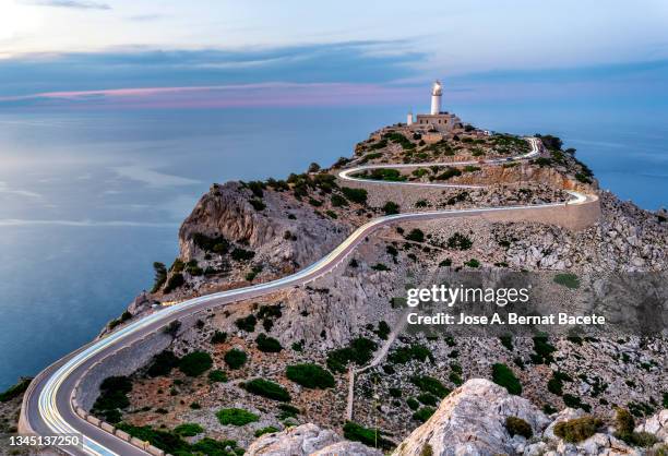 high angle view of light trails from cars on a road leading up to a lighthouse facing the sea at sunset. majorca island. - lighthouse mallorca stock pictures, royalty-free photos & images