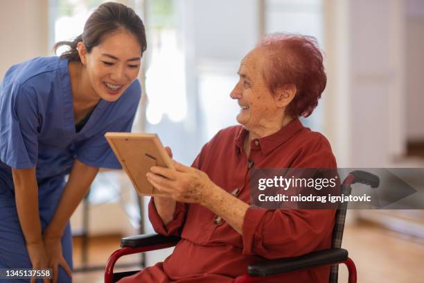 senior woman in wheelchair talking funny things from life while showing her photo to nurse at nursing home - paralysis stock pictures, royalty-free photos & images