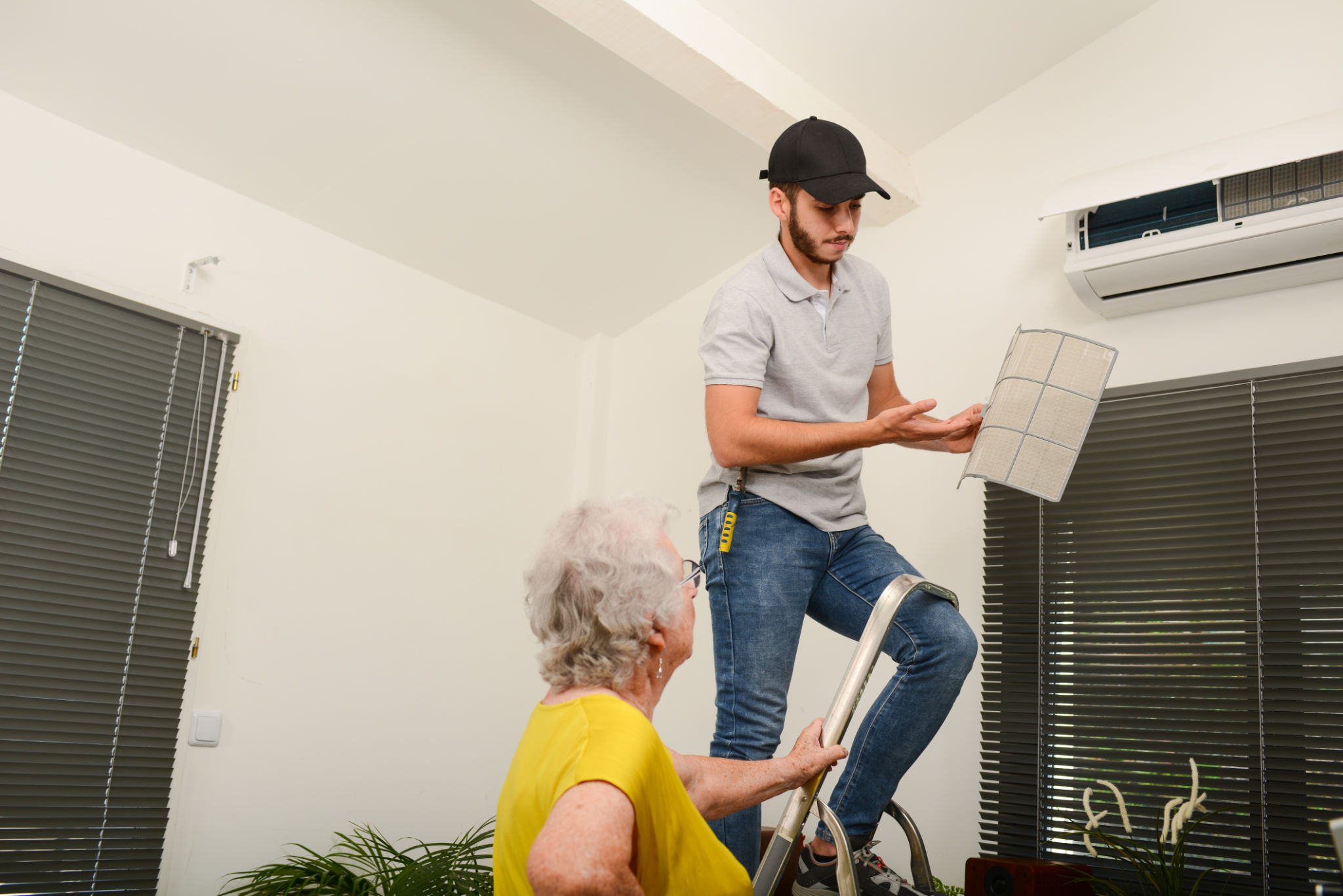 handsome young man electrician cleaning air filter on an indoor unit of air conditioning system in client house with senior woman handsome young man electrician cleaning air filter on an indoor unit of air conditioning system in client house with senior woman