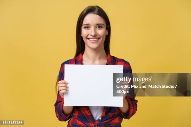 portrait of smiling young woman holding blank placard against yellow background - happy people holding a white board stock pictures, royalty-free photos & images