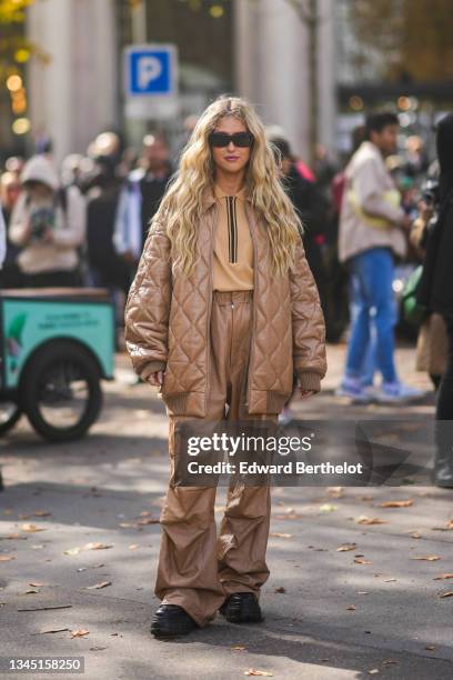 Emili Sindlev wears sunglasses, gold earrings, a beige pullover, a brown leather quilted / puffy oversized long coat from Miu Miu, matching brown...