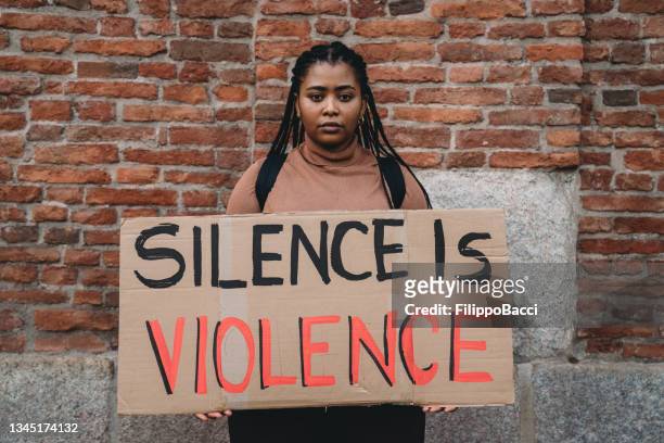 millennial-frau hält ein pappschild mit dem text "schweigen ist gewalt" darauf - mensch protest piktogramm stock-fotos und bilder