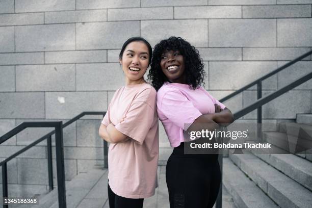 junge asiatische und afroamerikanische frauen posieren mit verschränkten armen - rücken an rücken stock-fotos und bilder