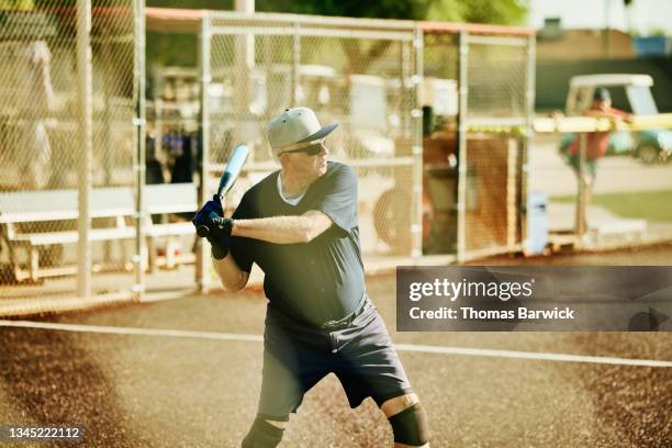 medium wide shot of senior softball player preparing to swing during at bat during game on summer morning - softball sport stock-fotos und bilder