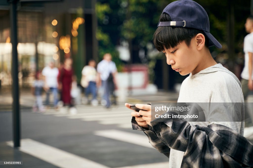 Asian teenage boy playing with cellphone outdoors on street