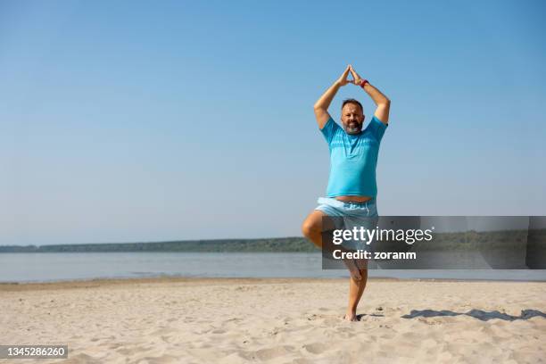man practicing yoga in tree pose on the beach on a clear day - fat man on beach stock pictures, royalty-free photos & images