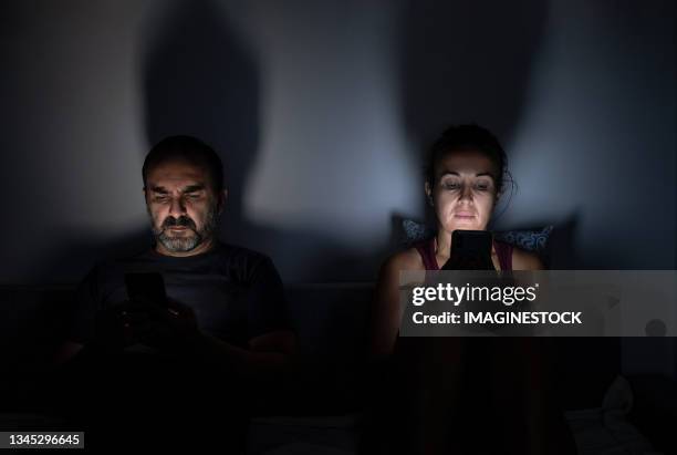 couple using their mobile phones on the sofa at home at night - couple couch imagens e fotografias de stock