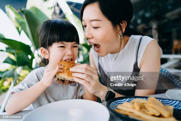 asian family having fun while enjoying lunch in an outdoor restaurant, mother and daughter are sharing and having a big bite eating a burger. family eating out lifestyle - kid eating restaurant stock pictures, royalty-free photos & images
