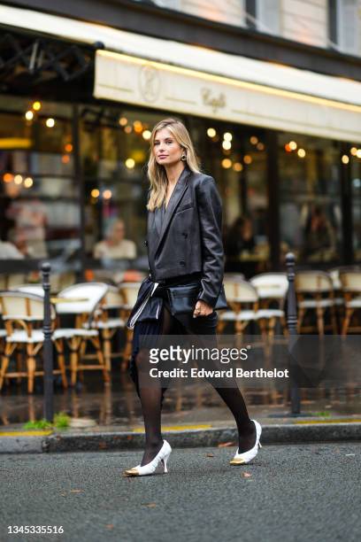 Xenia Adonts wears a black leather blazer jacket, a black leather clutch, a navy blue pleated skirt, tights, white pointed high heeled shoes with...