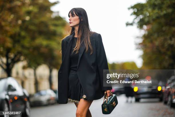 Leia Sfez wears a black oversized blazer jacket, a black ribbed top, a mini black skirt with attached jewelry, a green leather bag with golden...