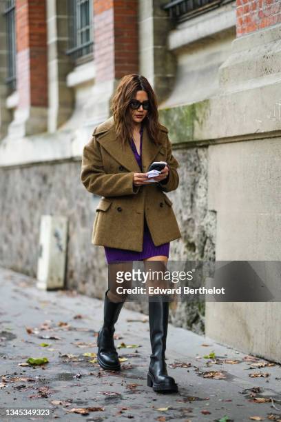 Christine Centenera wears sunglasses, a beige wool autumn jacket / coat with large lapels, a purple mini dress, black knee-high leather boots,...