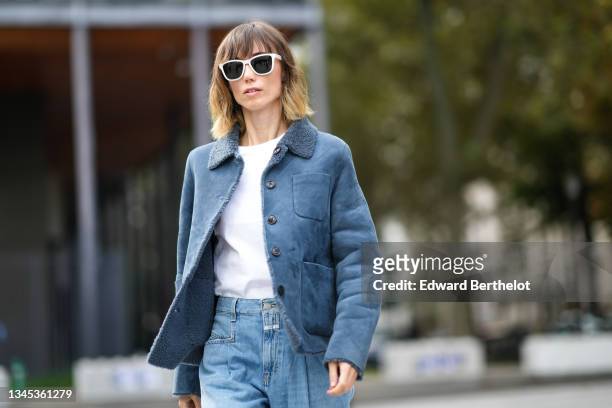 Anya Ziourova wears sunglasses, a white t-shirt, a blue suede autumn/winter jacket with fluffy inner wool lining, blue denim jeans pants, outside...
