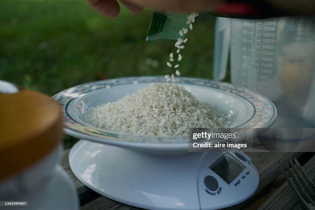 An amount of rice is being weighed over a white plate by using a digital scales. Some rice is putted onto a plate for weight measurements over a digital scale.