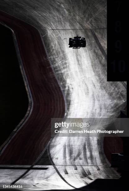 In back lit, high contrast light a Formula One car drives up the hill in the pit lane exit lane and on to the track next to the digital driver name...