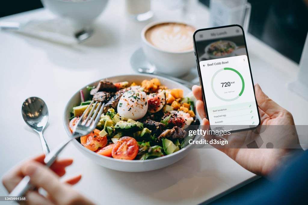 Close up of young woman using fitness plan mobile app on smartphone to tailor make her daily diet meal plan, checking the nutrition facts and calories intake of her beef cobb salad. Maintaining a balanced diet. Healthy eating lifestyle