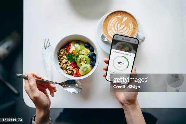 table top view of young woman using fitness plan mobile app on smartphone to tailor make her daily diet meal plan, checking the nutrition facts and calories intake of her breakfast fruit bowl. maintaining a balanced diet. healthy eating lifestyle - huntsman breakfast stock pictures, royalty-free photos & images