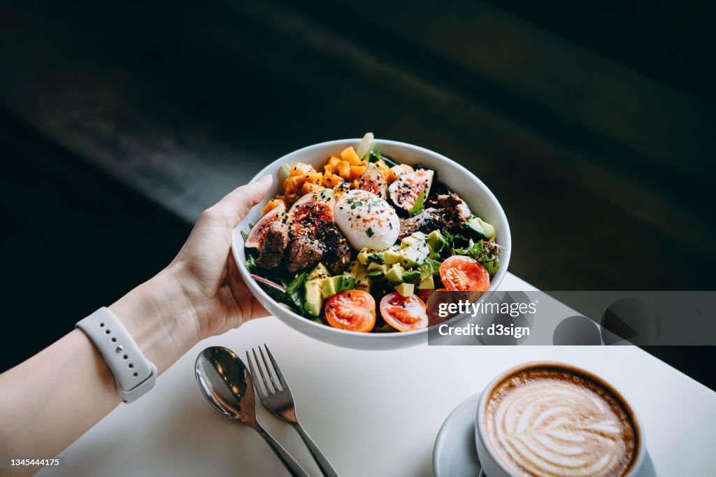 Close up of woman's hand holding a bowl of fresh beef cobb salad, serving on the dining table. Ready to enjoy her healthy and nutritious lunch with coffee. Maintaining a healthy and well-balanced diet. Healthy eating lifestyle