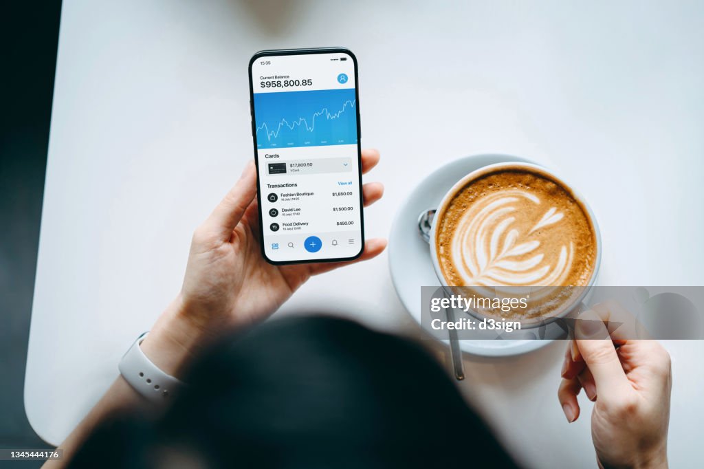 Overhead view of young woman enjoying a coffee break, managing finance and investment, checking financial stock market data on smartphone in cafe. Smart banking with technology