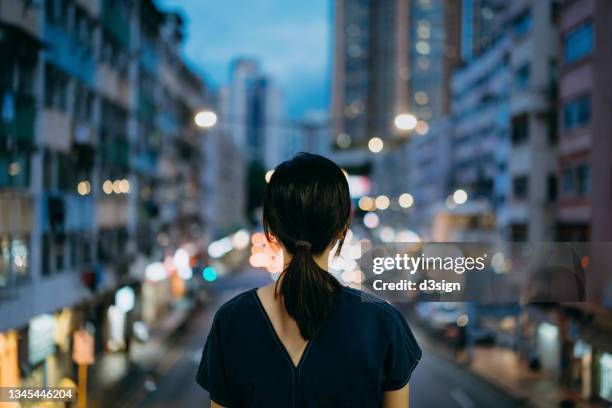 rear view of young asian woman in the city standing against illuminated city street at dusk - smart cities stockfoto's en -beelden