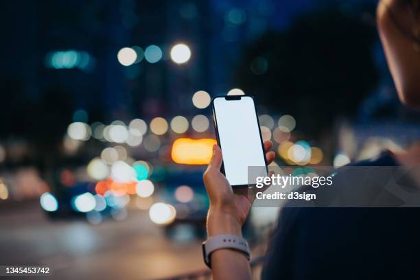 over the shoulder view of young woman using mobile app on smartphone to arrange taxi ride in downtown city street, with illuminated city traffic scene as background. smartphone with blank screen for design mockup - over the shoulder view stock-fotos und bilder