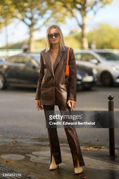 Guest wears sunglasses, a brown shiny leather blazer jacket, brown shiny leather slit / split flared pants, an orange shiny grained leather shoulder...