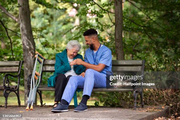 caregiver man with senior woman with crutches sitting on bench and greeting outdoors in park. - nursing assistant stock pictures, royalty-free photos & images