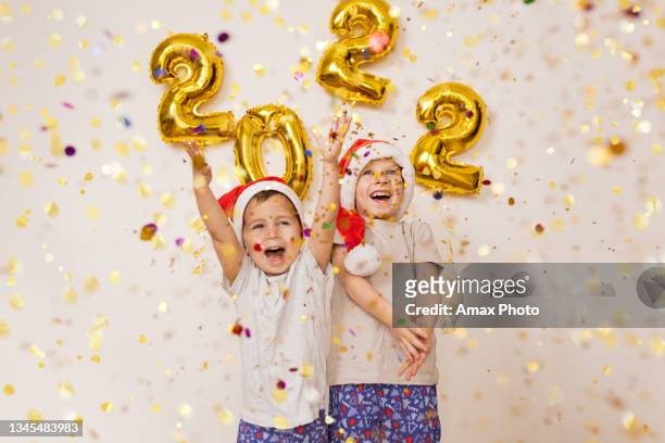 happy smiling kids in red santa hat are enjoying confetti near white wall with gold balloons. celebrating new year 2022 at home. christmas concept - 2022 a funny thing stock pictures, royalty-free photos & images