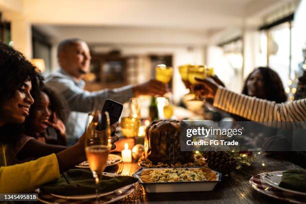 junge frau, die einen videoanruf macht / filmt / fotos von der familie macht, die zu hause auf das weihnachtsessen anstößt - african american christmas images stock-fotos und bilder