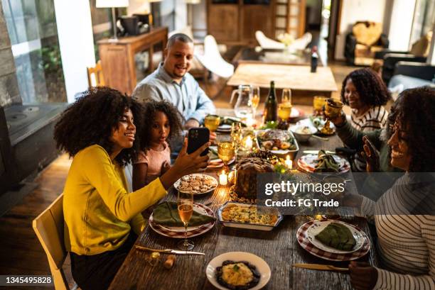 young woman doing a video call/filming/taking photos of the family toasting on christmas dinner at home - conference dining table stockfoto's en -beelden