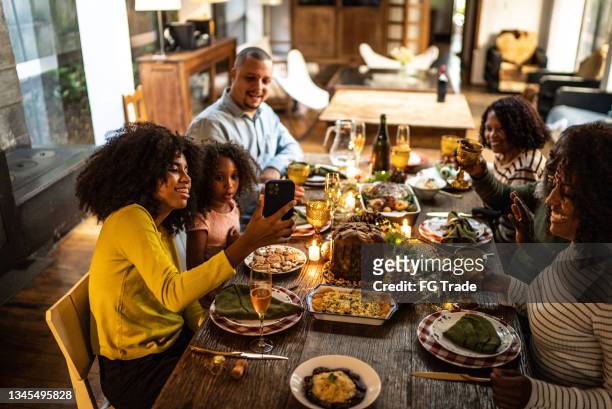 young woman doing a video call/filming/taking photos of the family toasting on christmas dinner at home - black family reunion stock pictures, royalty-free photos & images