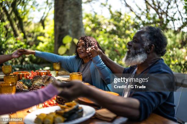 senior man saying grace before thanksgiving dinner at home - african at dining table stockfoto's en -beelden