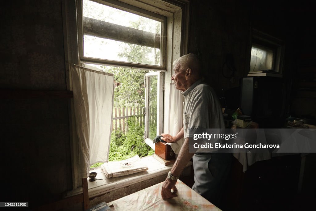 An old 97-year-old man stands at an open window in his house and looks into the distance.
