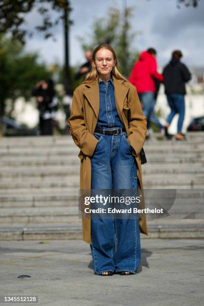 Alexandra Carl wears glasses, a brown long trench coat, a blue denim jumpsuit with flared pants, a black leather belt, outside Ludovic de Saint...