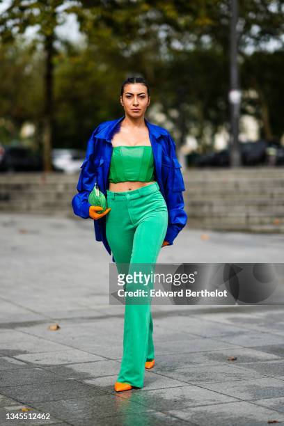 Guest wears a bold blue oversized jacket with pockets on the sleeves, a green corset cropped top, green high waist flared pants, orange pointed...