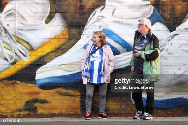 Fans of Colchester United stand beside an Adidas Trainer mural painted on the outside of the stadium by Artist Paul Curtis prior to the Sky Bet...