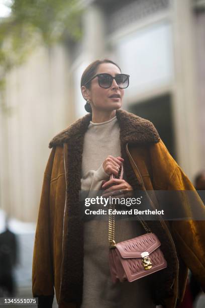 Geraldine Boublil wears sunglasses, earrings, a beige ribbed turtleneck wool long pullover dress, rings, a pink matte embossed leather handbag from...