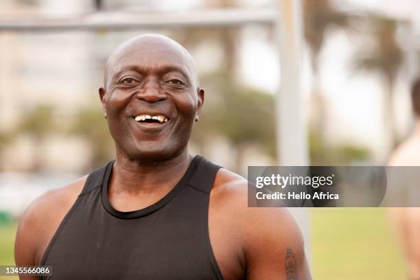 a strong happy smiling man showing his gold teeth and wearing a black sleeveless sports vest at an outdoor gym with lush green lawn visible in background at an urban city park - capped tooth stock-fotos und bilder
