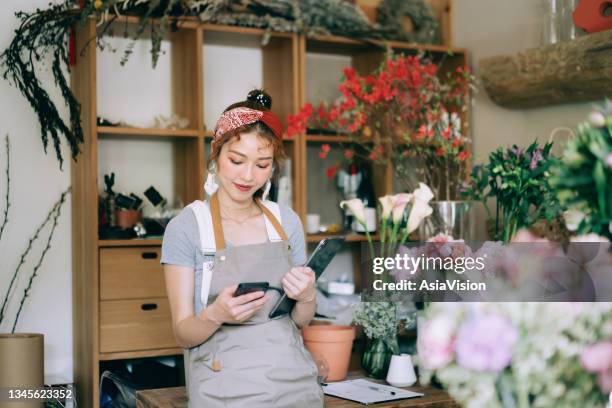 joven florista asiática, propietaria de una floristería de pequeñas empresas, que utiliza tabletas digitales y teléfonos inteligentes en el lugar de trabajo. comprobación de existencias, toma de pedidos de clientes, venta de productos en línea. rutin - asia lady selling flower fotografías e imágenes de stock