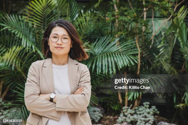 asian businesswoman standing with arms crossed in front of green plants - city life asia stock pictures, royalty-free photos & images