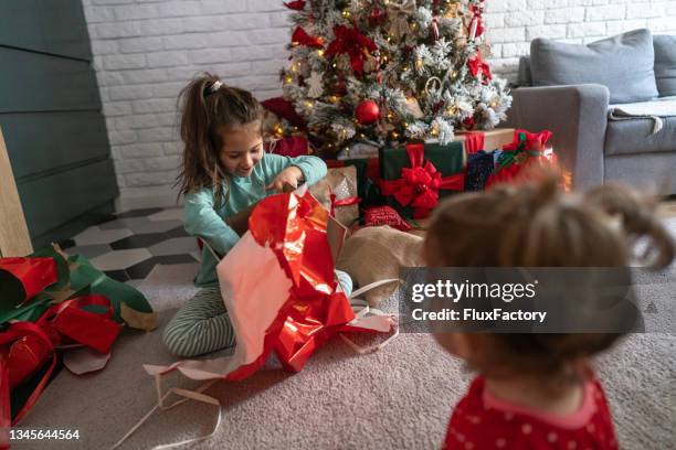 schönes mädchen kann ihr lächeln nicht verbergen, nachdem sie gesehen hat, was sie für ein weihnachtsgeschenk bekommen hat, während sie mit ihrer kleinen schwester ein geschenk auspackt - auspacken stock-fotos und bilder