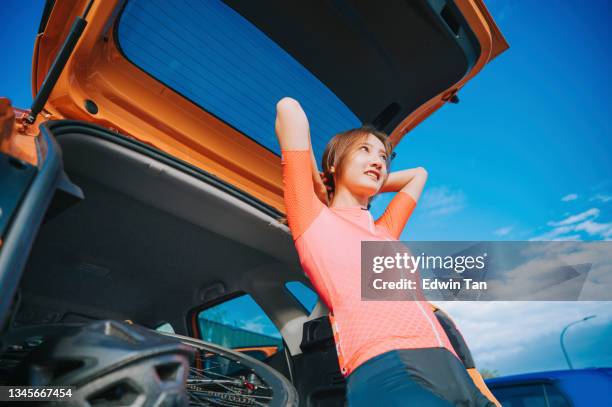 low angle view asian chinese young woman tying her hair getting ready for road bike trip adventure during weekend morning leaning on car trunk - camisola de ciclismo imagens e fotografias de stock