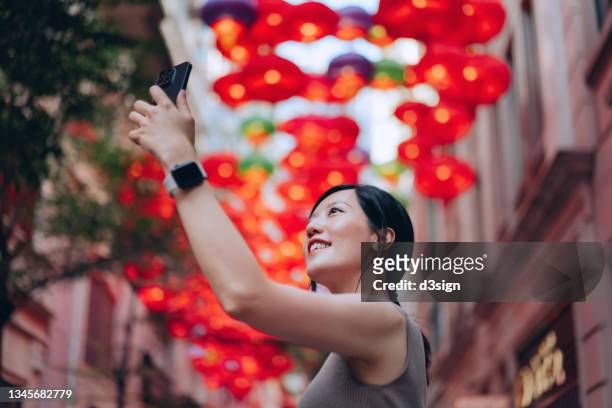 beautiful smiling young asian woman taking a selfie with smartphone in front of traditional chinese red lanterns hanging along the city street. traditional chinese culture, festival and celebration event theme - chinese festival stock-fotos und bilder