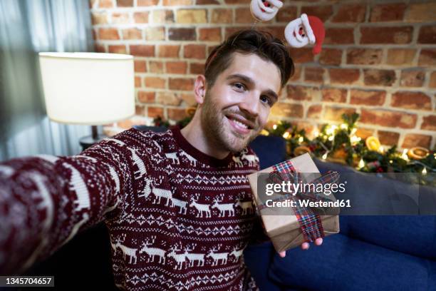 smiling man taking selfie with christmas present at home - weihnachtspullover mann stock-fotos und bilder