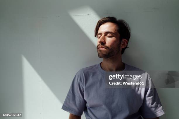 relaxed young man with eyes closed leaning on white wall - man closed eyes stock-fotos und bilder